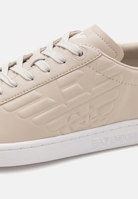 Béžová teniska na šněrování s vyraženým logem orla Emporio Armani na boku a bílou texturovanou podrážkou s označením EA7.
