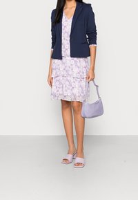 Námořnický blejzr přes fialové květinové šaty s vrstvenými volány, doplněné o fialové sandály na block podpatku a texturovanou kabelku přes rameno.