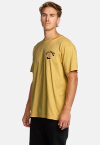 T-shirt di cotone giallo senape con maniche corte, dotata di un logo stampato in rosso e nero sul petto, vestibilità rilassata e scollo rotondo.