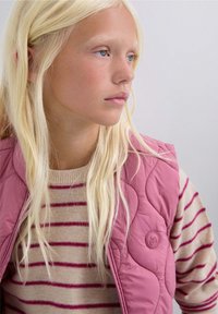 Chica rubia con piel clara y pecas, vistiendo un chaleco acolchado rosa sobre un suéter a rayas beige y rosa, mirando hacia la derecha.