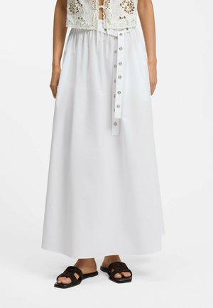 Frau trägt einen weißen, hoch taillierten Maxirock mit Schnappverschluss-Gürtel, kombiniert mit schwarzen flachen Sandalen und einem Häkelspitzen-Top.