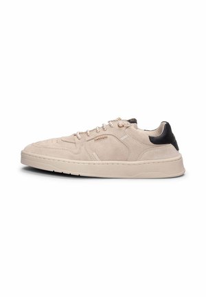 Zapatilla baja de ante beige con cordones blancos, lengüeta negra en el talón y suela de goma, mostrada de perfil sobre un fondo blanco.
