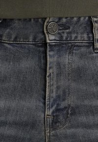 Jean en denim à lavage foncé avec un léger délavage, présentant une fermeture à bouton en métal et une taille côtelée en olive au sommet.