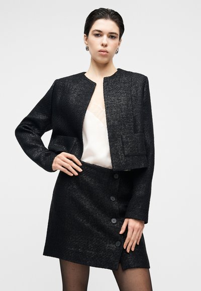 Giacca corta in tweed nero con collo rotondo, due tasche frontali, abbinata a una gonna a vita alta con chiusura laterale a bottoni.