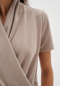 Gros plan sur une personne portant un haut beige à manches courtes de style cache-coeur avec un col plié et un tissu texturé.