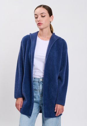 Vrouw draagt een blauwe pluizige open cardigan over een wit shirt en lichtblauwe spijkerbroek, staand tegen een effen lichte achtergrond.