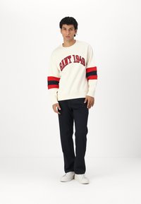 Kremowy sweatshirt z czerwonymi i granatowymi paskami na rękawach, z napisem "GANT 1949" w czerwonym kolorze. Połączony z ciemnogranatowymi spodniami i białymi sneakersami.