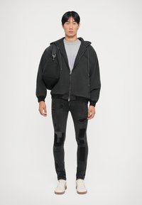Črna zip-up kapuca z zaobljenim ovratnikom, siva grafična majica, uničene črne kavbojke in bež superge. Teksturna tkanina in ozka prileganje.
