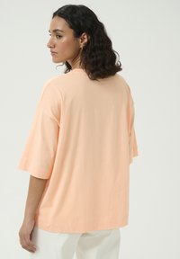 T-shirt à manches courtes, ample, en coton doux pêche. Présente un col rond et une silhouette décontractée. Détail de couture minimal au niveau de l'ourlet.