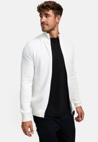 Chaqueta blanca de punto con un patrón texturizado, cuello alzado y cremallera completa, combinada con una camiseta negra y pantalones oscuros.