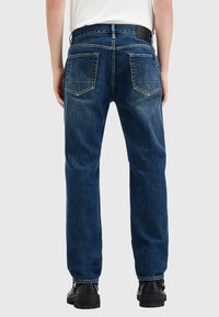 Vaqueros de denim azul oscuro con un corte relajado, que presentan dos bolsillos traseros y costuras visibles. Llevado con botas negras.
