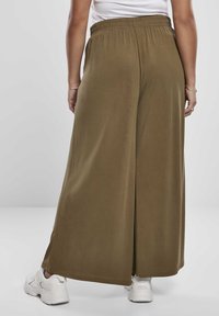 Personne portant un pantalon ample couleur vert olive et des baskets blanches, vue de dos sur un fond gris uni.