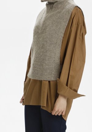 Villainen harmaa hihaton neuleliivi, joka on päällä löysän ruskean pitkähihaisen paidan päällä, jossa on lasketut olkapäät ja tummat housut seisovalla henkilöllä.