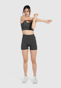 Top deportivo negro corto y pantalones cortos de talle alto hechos de una tela elástica, con una textura suave. La modelo está estirándose con zapatillas blancas.