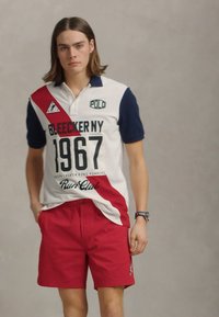 Polo majica v beli, mornarsko modri in rdeči barvi s tiskom "BLEECKER NY 1967" in kratkimi rdečimi hlačami. Vsebuje kontrastne barvne bloke in logotipne akcentne detajle.