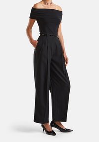 Černý top s odkrytými rameny v kombinaci s černými kalhotami s vysokým pasem a širokými nohavicemi, doplněný lesklými černými podpatky a tenkým černým páskem.