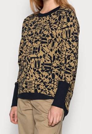 Femme portant un haut à manches longues marron et noir avec un motif abstrait, des poignets noirs et un pantalon beige, vue du cou aux hanches.