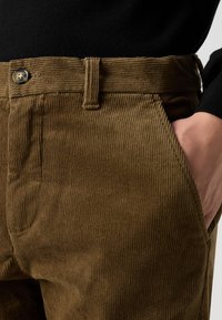 Hnědé manšestrové kalhoty s vertikálním žebrováním, zapínáním na knoflíky v pase a bočními kapsami. Hladká textura s klasickým, na míru šitým designem.