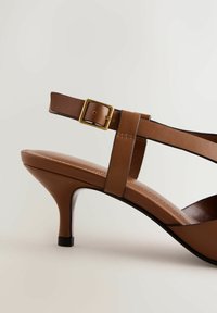 Brūna augstpapēžu sandale ar šauru potītes siksniņu, zelta sprādzes detaļu un gludu ādas tekstūru. Raksturīga pirksta daļa ar smailu formu un strukturēts papēdis.