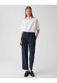 Witte katoenen blouse met poffende mouwen, gecombineerd met marineblauwe geplooide broeken. Model draagt zwarte instappers en staat tegen een lichte achtergrond.