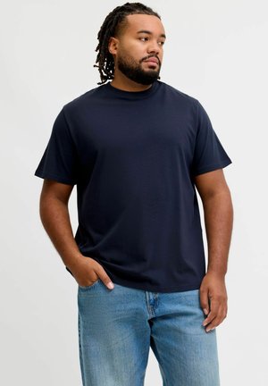 Mand med dreadlocks iført en mørkeblå t-shirt og lyseblå jeans, stående med den ene hånd i lommen mod en ensfarvet baggrund.
