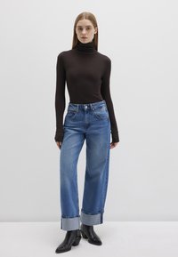 Brown turtleneck top, high-waisted blue jeans with rolled cuffs, black heeled boots, model standing against a plain white background.
