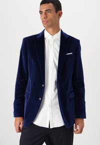Marineblauwe fluwelen blazer met een notch-lapel, twee voorkruiszakken en zilveren knopen, gecombineerd met een wit overhemd eronder.