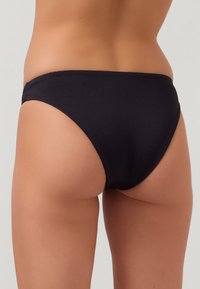 Eine Frau trägt schwarze Bikinihosen, von hinten gezeigt, vor einem neutralen hellen Hintergrund.