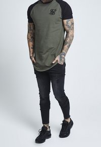 Olivgrön t-shirt med svarta raglanärmar, prydd med en liten svart logotyp på bröstet. Bärs med slitna svarta skinny jeans och svarta sneakers.