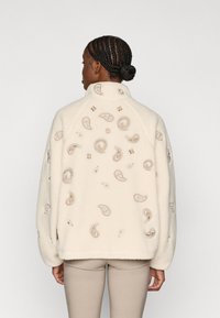 Κρέμα φλις ζακέτα με ψηλό γιακά, διακοσμημένη με κεντημένα σχέδια paisley σε μπεζ χρώμα και υπερβολικά μανίκια. Λείος υφασμάτινος χαρακτήρας, χαλαρή εφαρμογή.