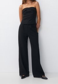 Haut noir sans manches avec des coutures à l'avant, associé à un pantalon noir à jambes larges. Le modèle porte des chaussures noires pointues avec des clous. Texture de tissu lisse.