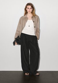 Ung kvinde iført beige jakke, hvid top, brede sorte bukser, sorte sandaler, holder en sort vævet clutch, står op ad en ensfarvet væg.