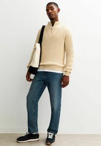 Pullover beige a costine con colletto a mezza zip, abbinato a jeans blu a gamba dritta e sneaker nere. Gli accessori includono una borsa bianca e nera.