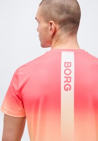 Άνδρας με κοντά μαλλιά φοράει αθλητική μπλούζα με βαθμιαία αλλαγή χρώματος από ροζ σε πορτοκαλί, με κατακόρυφη λευκή ρίγα και κόκκινο κείμενο «BORG» στην πλάτη.