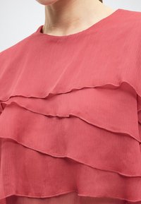 Κοντινό πλάνο ενός ατόμου που φοράει ροζ μπλούζα με στρώσεις και βολάν, με λεπτή κάθετη υφή και στρογγυλή λαιμόκοψη.