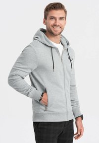 Hellgraue Zip-Hoodie mit verstellbarem Kapuzenzug, Seitentaschen und gerippten Bündchen. Mit kleinem Logo auf der Brust. Weicher Stoff.