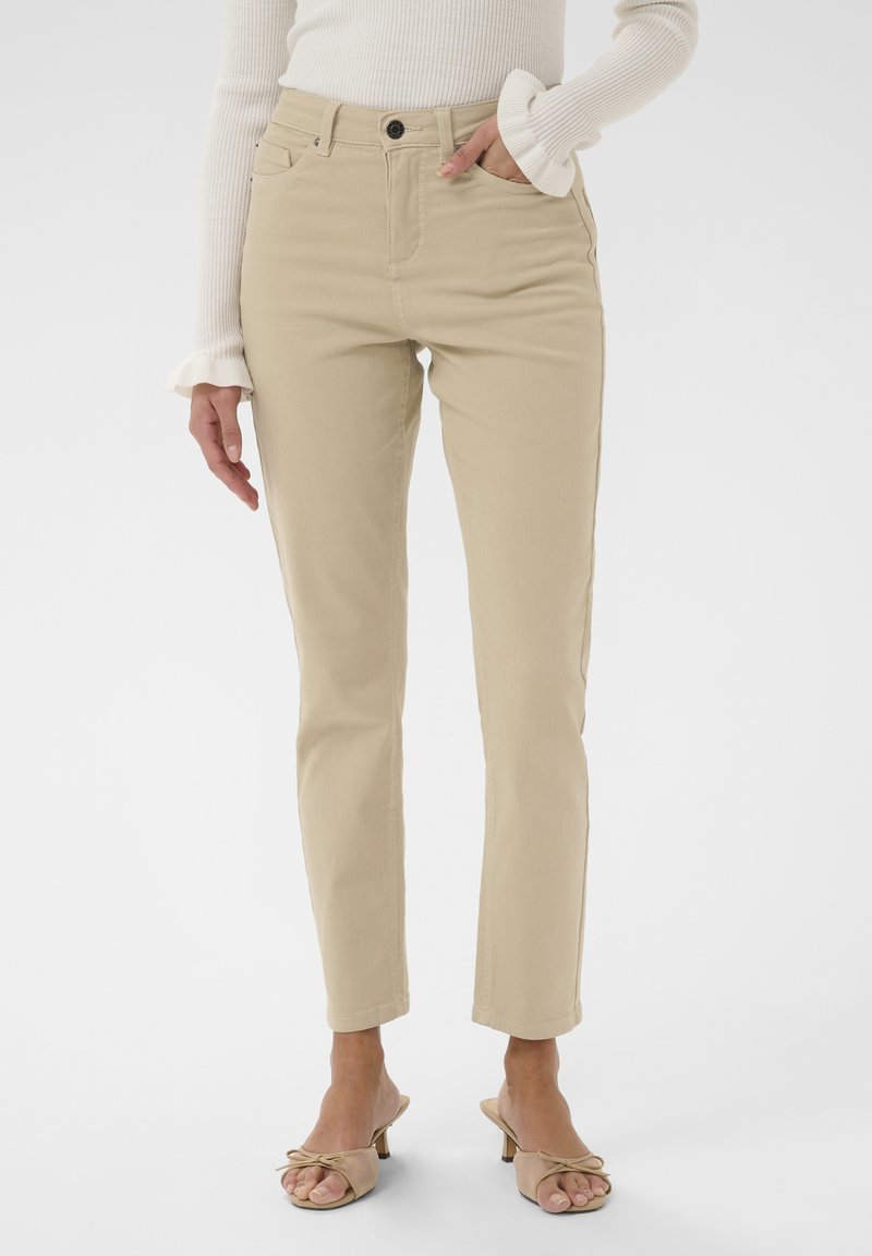 Femme portant un pantalon beige moulant, un pull crème côtelé avec des manches à volants, et des sandales à talons ouvertes beiges avec des nœuds.