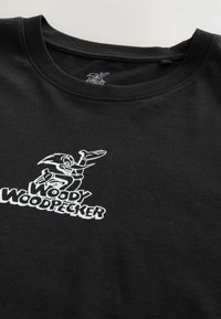 Čierne bavlnené tričko s krátkymi rukávmi, na prednej strane s bielou grafikou Woodyho Woodpeckera a textom v tučnom písmovom štýle.