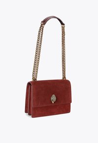 Sac à main en daim rouge avec une chaîne en métal doré et une poignée en cuir. Il présente un fermoir décoratif en forme de coquillage sur un rabat.