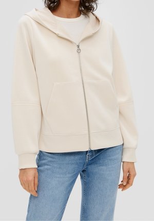 Persona che indossa una felpa con cappuccio color crema con cerniera e tasche frontali, e jeans azzurri, in piedi contro uno sfondo semplice.