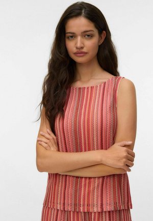 Jonge vrouw met lang donker haar die een mouwloze rood-roze gestreepte top draagt, armen gekruist, tegen een effen witte achtergrond.