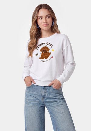 Ung kvinde med bølget hår iført en hvid sweatshirt med et brunt hvalpebillede og tekst "Puppy Girl", kombineret med lyseblå jeans.