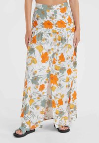 Femme portant une jupe maxi blanche avec un imprimé floral orange, jaune et vert et des boutons sur le devant, accompagnée de sandales tressées bleues.