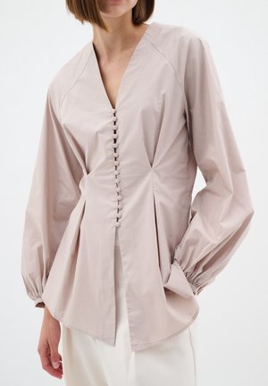 Vrouw draagt een lichtbeige blouse met lange opgeblazen mouwen en een rij bedekte knopen aan de voorkant, gecombineerd met witte broek.