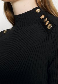 Pull noir côtelé avec un col haut et cinq boutons dorés à l'épaule, offrant une apparence texturée et une silhouette structurée.