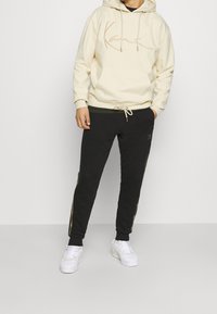 Krémová mikina s kapucí a přední kapsou, vyšitým vzorem, v kombinaci s černými tepláky se zlatými bočními pruhy a bílými teniskami.