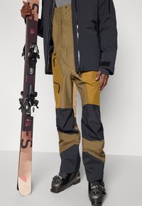 Férfi fekete síkabátban, több színű barna és fekete sínadrágban, fekete sícipőben, aki egy pár barna sílécet tart, fekete kötésekkel.