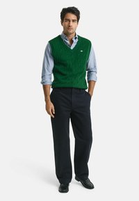 Groene gebreide mouwloze trui met een kabelpatroon, gedragen over een blauw-wit gestreept shirt, gecombineerd met donkere broeken en zwarte schoenen.