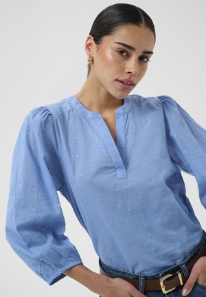 Jeune femme aux cheveux foncés portant des boucles d'oreilles créoles dorées, un chemisier bleu clair texturé et une ceinture marron avec une boucle dorée, posant devant un fond blanc.