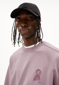 Sweat-shirt en tissu lavande clair avec un logo en relief sur la poitrine ; associé à une casquette noire avec un design de lettres brodé.